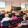 Auf dem Bild sitzen viele Menschen auf Stühlen mit dem Rücken zur Kamera. Vorne ist eine große Leinwand, darauf liest man: Betreuungsrechtsreform. Eine Frau steht vor den Zuhörerinnen auf den Stühlen und hält ein Mikrofon in der Hand.