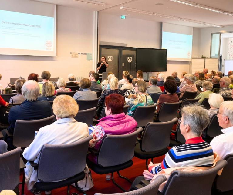 Auf dem Bild sitzen viele Menschen auf Stühlen mit dem Rücken zur Kamera. Vorne ist eine große Leinwand, darauf liest man: Betreuungsrechtsreform. Eine Frau steht vor den Zuhörerinnen auf den Stühlen und hält ein Mikrofon in der Hand.