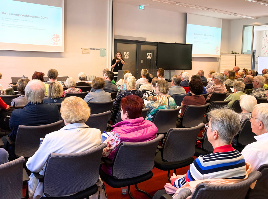 Auf dem Bild sitzen viele Menschen auf Stühlen mit dem Rücken zur Kamera. Vorne ist eine große Leinwand, darauf liest man: Betreuungsrechtsreform. Eine Frau steht vor den Zuhörerinnen auf den Stühlen und hält ein Mikrofon in der Hand.