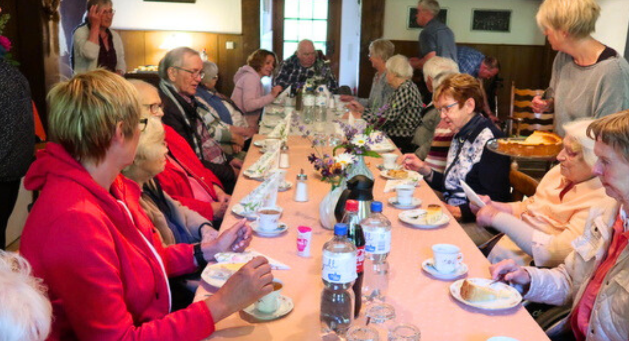 Eine Gruppe älterer Menschen sitzt an einem langen Tisch auf dem Kaffee und Kuchen steht.