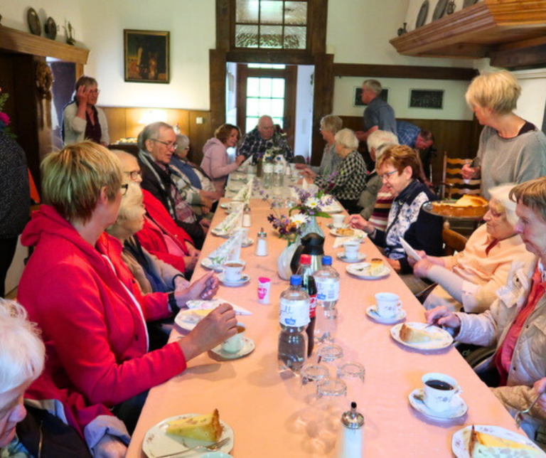 Eine Gruppe älterer Menschen sitzt an einem langen Tisch auf dem Kaffee und Kuchen steht.